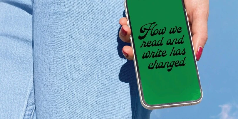 "Hand holding smartphone with green screen displaying 'How we read and write has changed', with denim jeans in background.